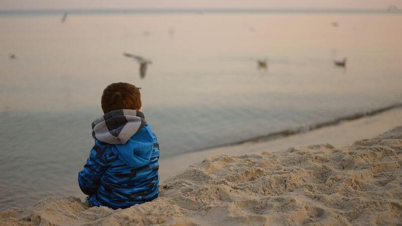 dziecko w niebieskiej kurtce siedzące samotnie na plaży. W oddali widać mewy